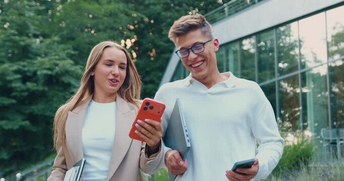 Two young adults man and woman strolling in a park while checking their mobile phones, sharing something on the screen and smiling during a friendly conversation