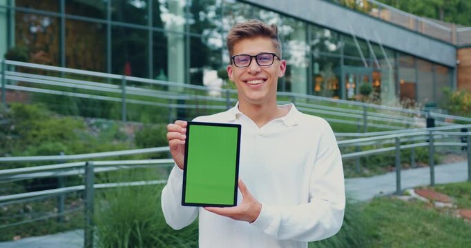 Young man smiling and presenting tablet with green screen in landscaped urban campus setting, front view, daylight exposure, casual white shirt and glasses, confident promotional pose