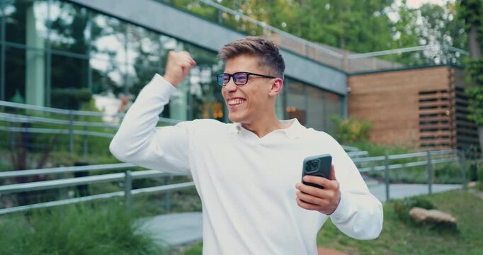 Attractive young man wearing white shirt holding phone and celebrating success, looking at cellphone screen, raising fist with excitement at modern glass building background outdoors
