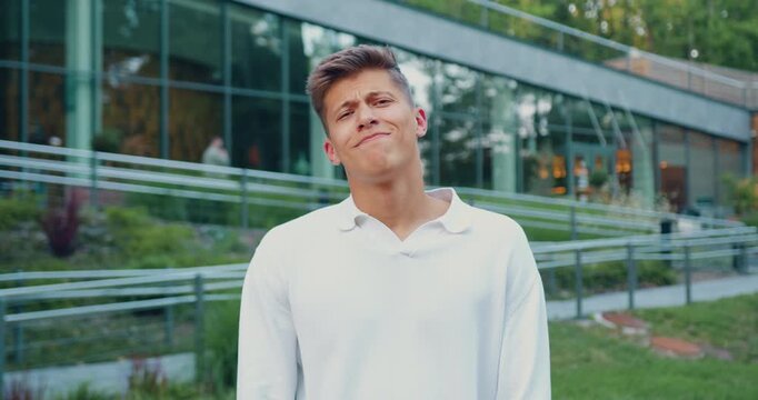 Young man outdoors in front of glass building showing surprised then amused facial reactions with subtle head tilt and shoulder movement, casual white shirt, daylight portrait scene