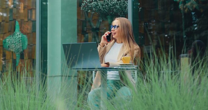 Busy businesswoman sitting on an outdoor terrace during lunch break, talking on smartphone, drinking coffee, and typing on laptop. Professional manager focused on remote work, multitasking, and modern