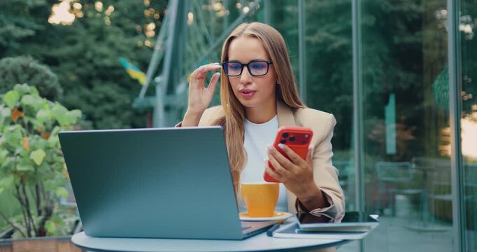 Young professional woman checking smartphone while working on laptop at outdoor cafe under soft daylight, thoughtful expression and casual business attire, modern terrace setting with greenery