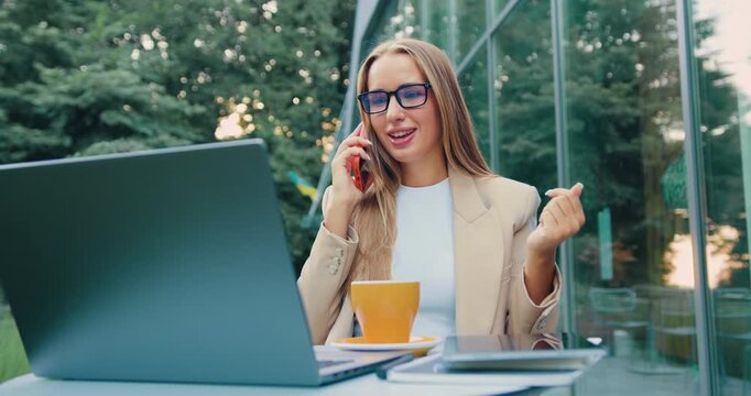 Young professional woman talking on smartphone and gesturing at outdoor cafe with laptop and coffee in daylight scene - energetic remote work moment in urban setting