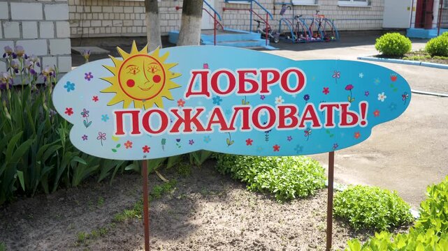 A bright blue welcome sign with a smiling sun illustration and red Russian text stands on green grass in front of a kindergarten yard with playground equipment.