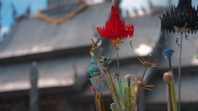 red paper ornaments and thai baht banknotes on sticks for buddhist donation, colorful crafted decorations with money attached near blurred shrine, Wat Sri Suphan Silver Temple