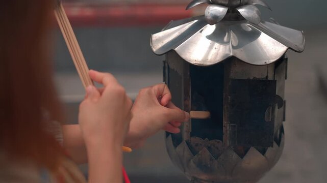 woman lighting incense sticks at metal temple burner, female hands holding spiritual offering near silver fire container for buddhist prayer 4k