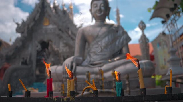 burning colorful candles in front of blurred silver buddha statue, lit fire offerings near large metallic religious sculpture outside 4k