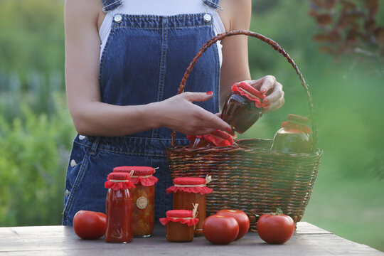 Woman arranging homemade tomato products in jars and glass bottles into wicker basket, preparing for farmers market, farm to table small business concept