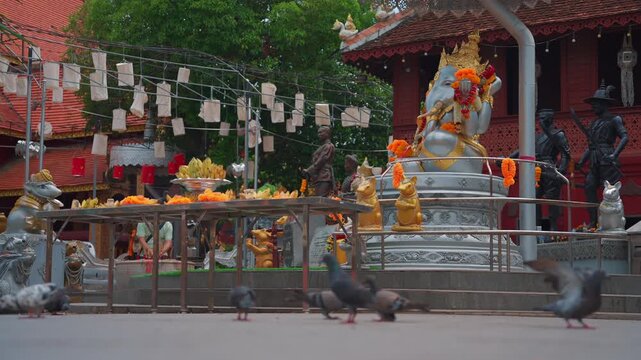 silver ganesha statue adorned with marigold garlands at thai temple, pigeons walking near hindu elephant deity shrine featuring fruit offerings 4k