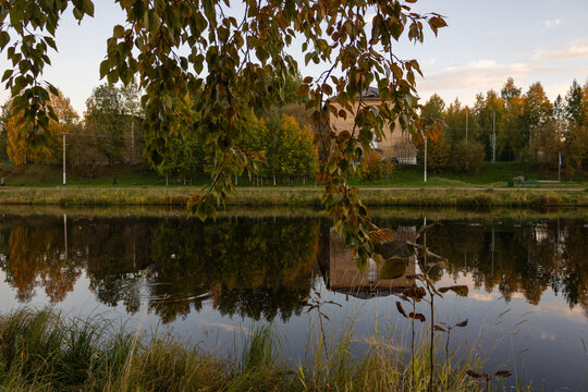 Peaceful panorama of an autumn city pond reflecting a brick school building and golden trees through hanging birch branches in Russia.
