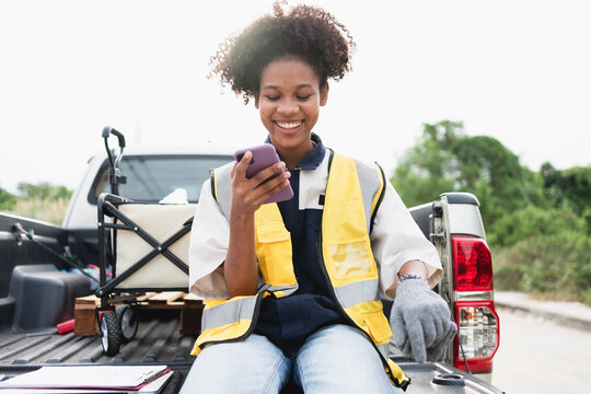 Smiling female field worker wearing a high visibility safety vest and gloves while using a smartphone beside a utility truck, representing mobile communication, outdoor work, and safety.