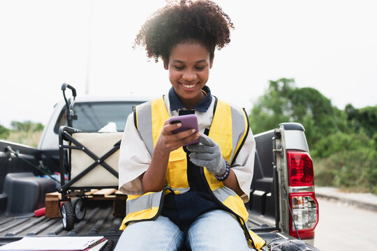 Smiling female field worker wearing a high visibility safety vest and gloves while using a smartphone beside a utility truck, representing mobile communication, outdoor work, and safety.