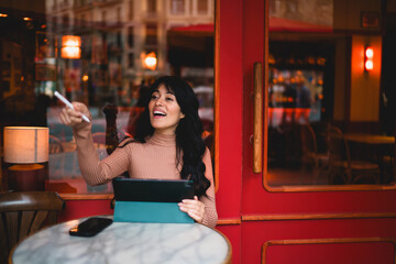 Excited Caucasian female student pointing stylus pen forward while holding digital tablet at cafe terrace table, discovering e-commerce flash sale discount deals and retail app offers © BullRun