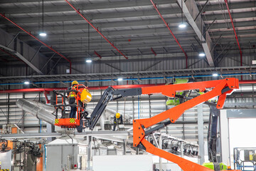 Workers wearing safety equipment working on an orange boom lift for ceiling maintenance inside a large modern factory. © Rangsarit
