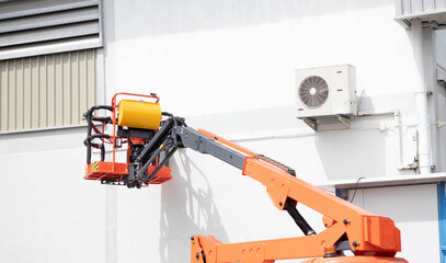 Orange boom lift aerial work platform elevated near air conditioner condenser unit on building wall for maintenance and repair work. © Rangsarit