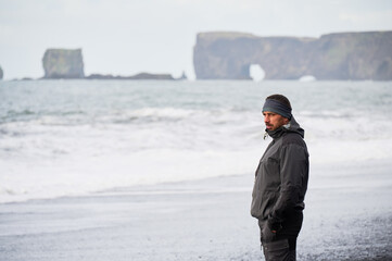 Male tourist stands beside breaking waves on Reynisfjara black beach with Dyrholaey natural arch in distance, framed with open space for travel, Iceland tourism and rugged coastal lifestyle themes © anatoliy_gleb