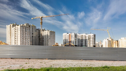 Apartment buildings and cranes stand at a construction site behind a long metal fence, with bare ground in the foreground during daytime © Anoo