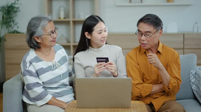 Happy Asian young woman holding credit card shopping online with her senior parents using a laptop computer on a sofa at home, family e-commerce digital finance and technology lifestyle concept.