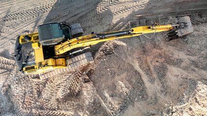 An excavator at a construction site reaches across churned ground with its boom and bucket while tracked treads mark the sandy surface © Anoo