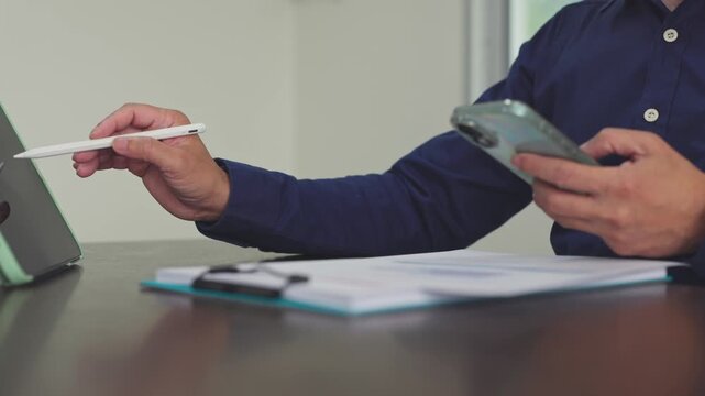 Businessman uses digital tablet pen mobile phone review paperwork at office desk. Corporate financial audit analysis, electronic document signature verification, accounting budget report.