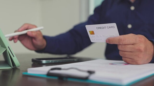 Man holds credit card while examining financial paperwork beside digital tablet in office. Digital payment verification, electronic commerce transaction, corporate accounting audit.
