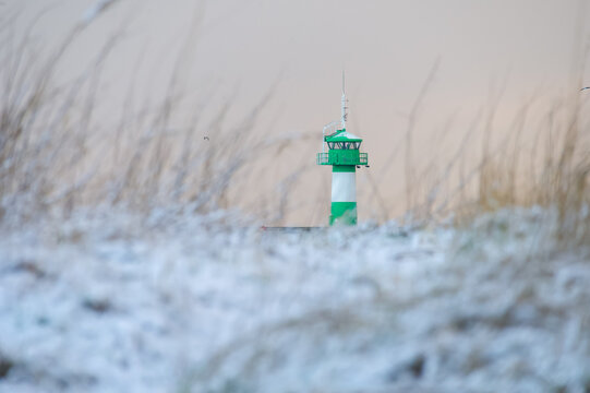 Gr&uuml;n-wei&szlig;er Leuchtturm in Travem&uuml;nde steht hinter verschneiten D&uuml;nen und Halmen bei sanftem, hellem Winterlicht an einer verschneiten K&uuml;stenlandschaft.