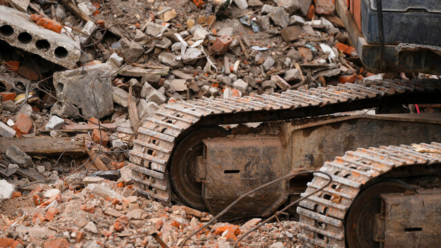 Steel crawler tracks and undercarriage of an excavator resting on broken bricks and concrete rubble at a demolition site, showing heavy equipment details