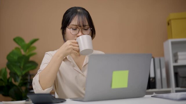 Young Asian woman wearing glasses working at laptop using digital pen for design or note-taking in modern home office