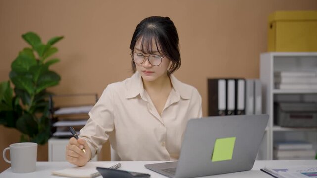 Young woman with glasses working on laptop at desk in modern office with plant and shelves in background