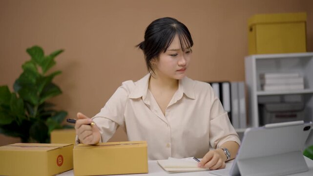 Young woman writing notes and preparing cardboard box for delivery in cozy office with plants and shelves