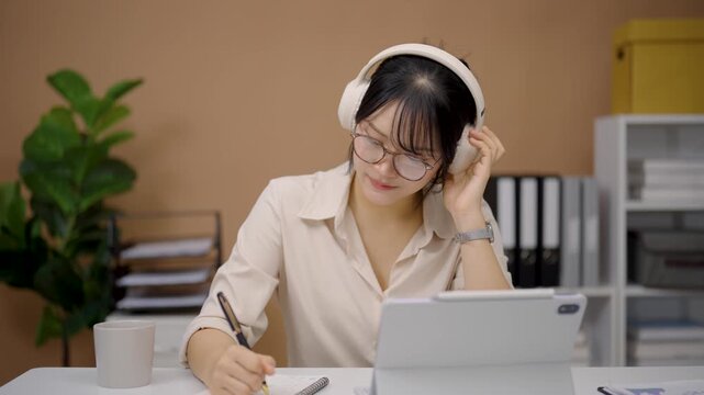 Young woman wearing headphones working on tablet computer taking notes in modern home office environment with plant and organizer