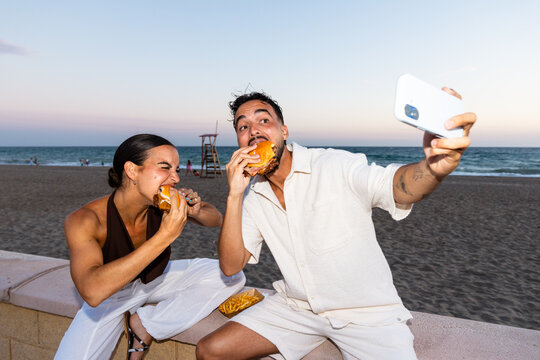 Happy couple enjoying burgers taking selfie on beach