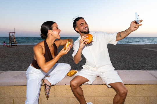 Happy couple eating burgers taking selfie on beach