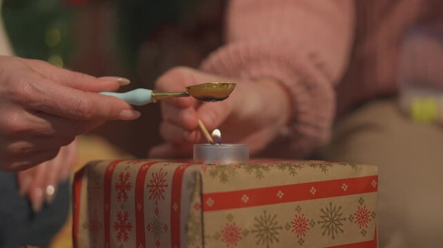 Detailed holiday footage captures hands as they melt sealing wax in a metal spoon above a small candle flame. Macro process view presents traditional gift decoration with strong commercial appeal.