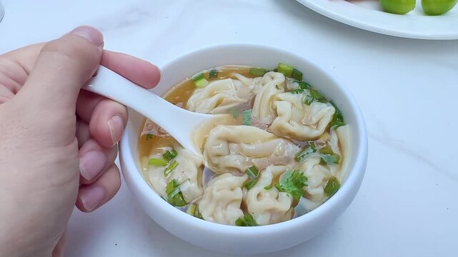 Hand Holding Spoon Over Bowl of Wonton Soup on Asian Restaurant Table