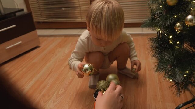 At Christmas, a toddler boy sorts gold ornaments on the floor beside the tree. The top down view highlights organization skills, festive play and seasonal joy for lifestyle media.