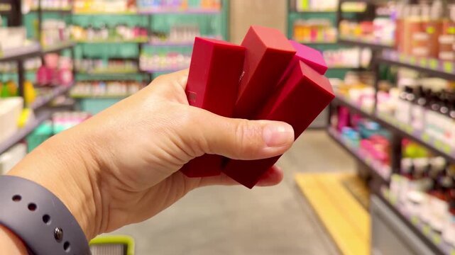 Hand Holding Several Colorful Lipsticks in Cosmetics Store &ndash; Choosing Between Different Shades Against Makeup Shelves