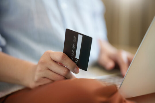 Close-up of a woman holding a black payment card while using a laptop at home. The scene captures online shopping, digital transactions and convenient cashless payment in everyday life.