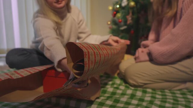 Warm lifestyle footage shows two women who measure patterned wrapping paper around a Christmas present. Medium floor view conveys gift wrapping preparation in a polished lifestyle scene.