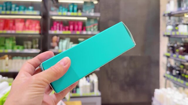 Hand Holding Shampoo Bottle and Hair Care Box in Cosmetics Store &ndash; Hair Oil, Tinted Balm and Conditioner Against Hair Care Shelves