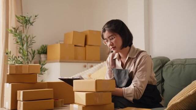 Asian female small business owner holding clipboard file and pen, inspecting customer order list carefully behind stacked parcel boxes.