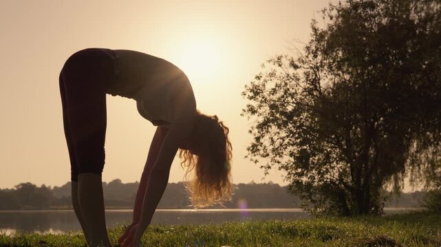 A young woman performs a forward bend yoga pose on grass near a lake at sunrise, embracing mindfulness and flexibility. The serene environment highlights relaxation and healthy lifestyle