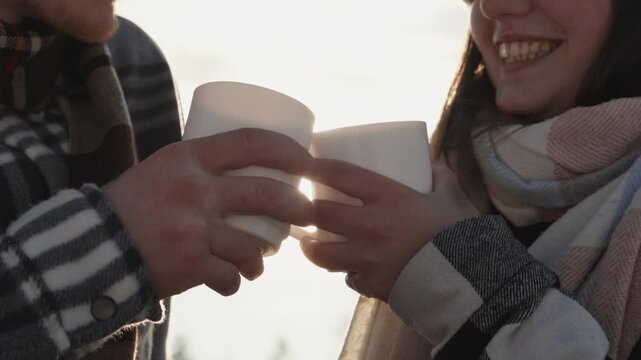 A young couple enjoys a hot beverage outdoors, dressed in striped and plaid scarves, celebrating a tender winter moment. Their smiles and gentle touch evoke warmth and connection