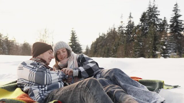 A loving couple spends quality time lying on a blanket in the snow, surrounded by pine trees and winter scenery. Their warm clothing and closeness evoke comfort and romance