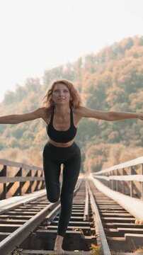 Athletic female performs yoga balance pose on abandoned railway bridge surrounded by golden autumn trees. Tranquil outdoor setting highlights harmony with nature and mindfulness