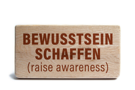 Holzschild mit Gravur &bdquo;Bewusstsein schaffen (raise awareness)&ldquo; auf wei&szlig;em Hintergrund
