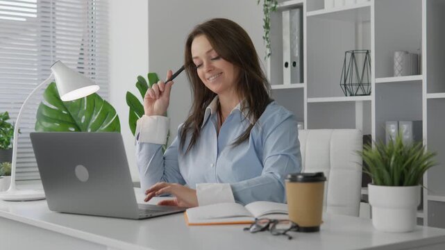 Focused female entrepreneur sits at a stylish white desk, writing in a notebook while using her laptop. Contemporary office environment with natural light and trendy decor elements