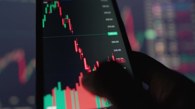 A hand holds a smartphone showing a dynamic candlestick chart, reflecting real time trading activity and market fluctuations. The blurred background features additional financial graphs and numbers