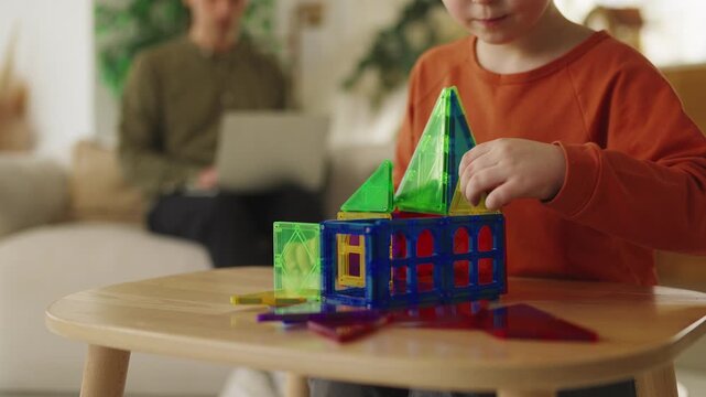 Little boy plays with colorful magnetic toy blocks while dad works nearby