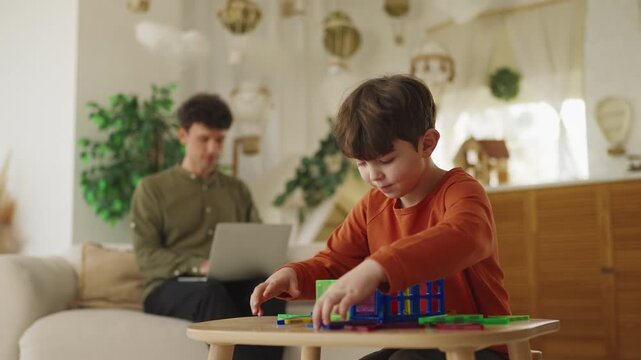 Little boy assembles toy building blocks while his father works on laptop
